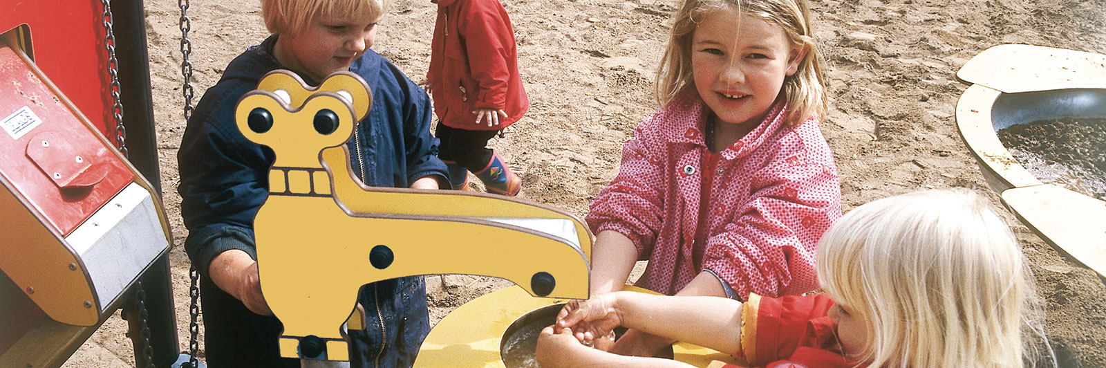 Kinder spielen an einer Wasserspielstation auf einem Spielplatz, ein Mädchen lächelt und schaut in die Kamera.